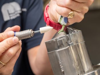 Hand-held rotary tool with a white pointed mounted felt beside diamond compound being applied to a metal workpiece