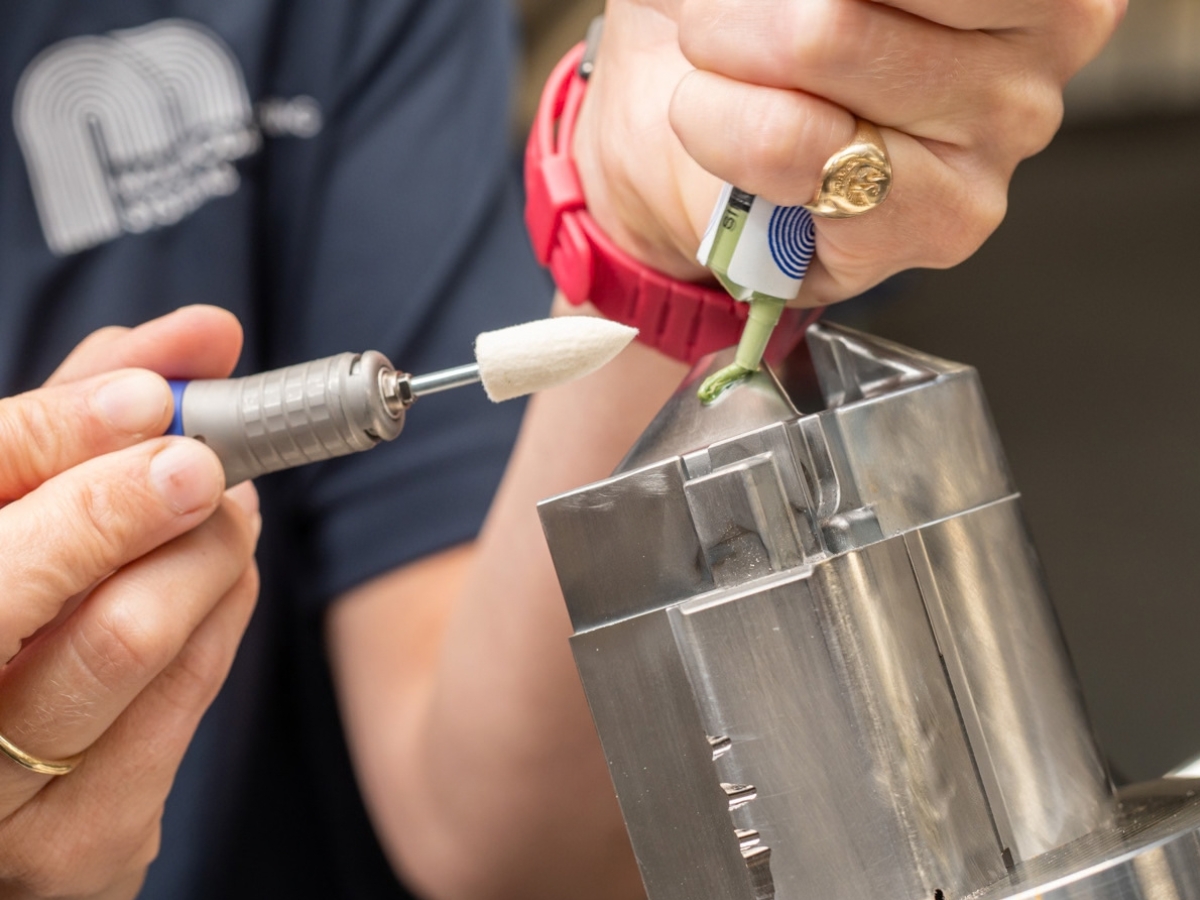 Hand-held rotary tool with a white pointed mounted felt beside diamond compound being applied to a metal workpiece